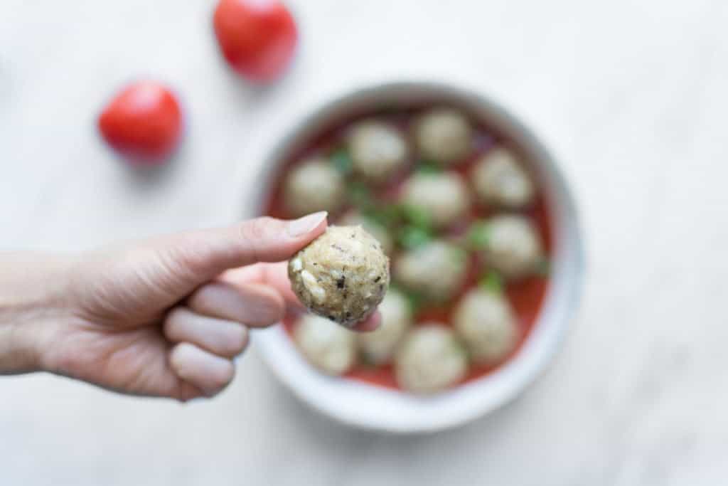 A close up of a chicken meatball in front of a blurred photo of a dish of meatballs in a tomato sauce.
