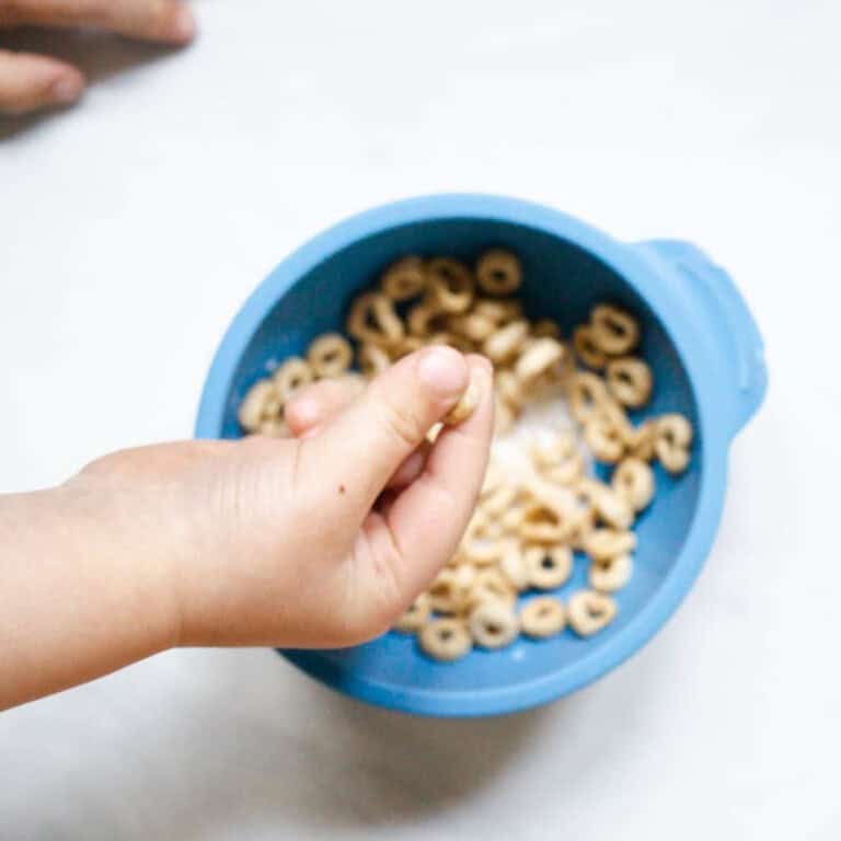 baby using pincer grasp to pick up wet cheerios from a blue bumpkins bowl