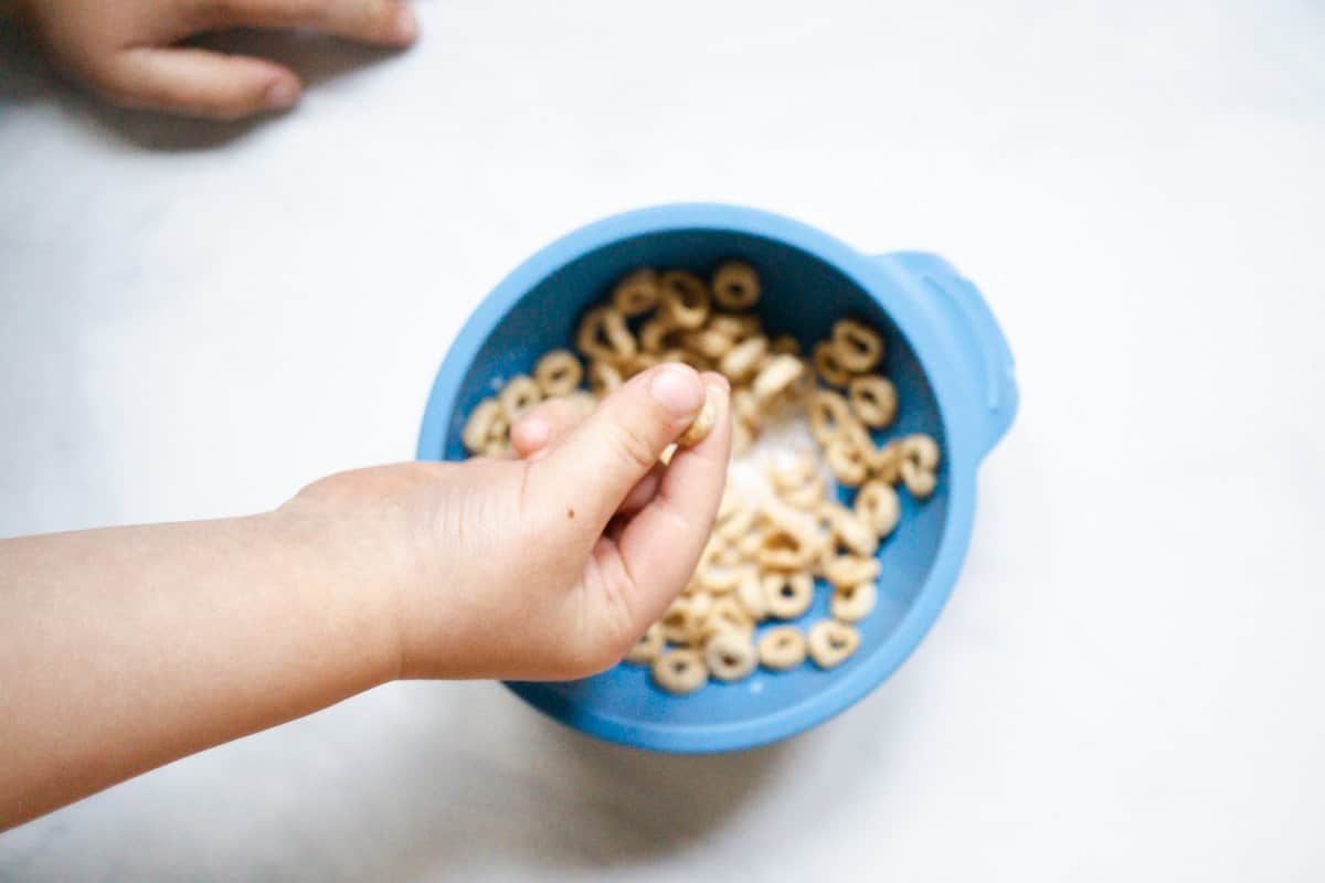 baby using pincer grasp to pick up wet cheerios from a blue bumpkins bowl
