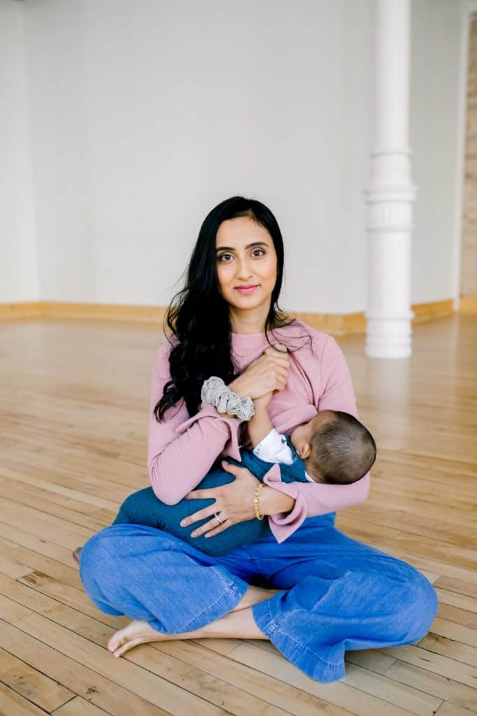 Mother with long black hair sits cross legged on a hard wood floor nursing a baby.