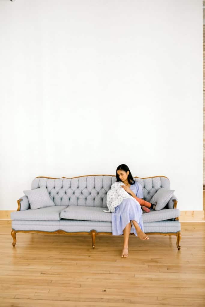 Mother with long black hair nurses baby under a blanket on a couch in front of a white wall.