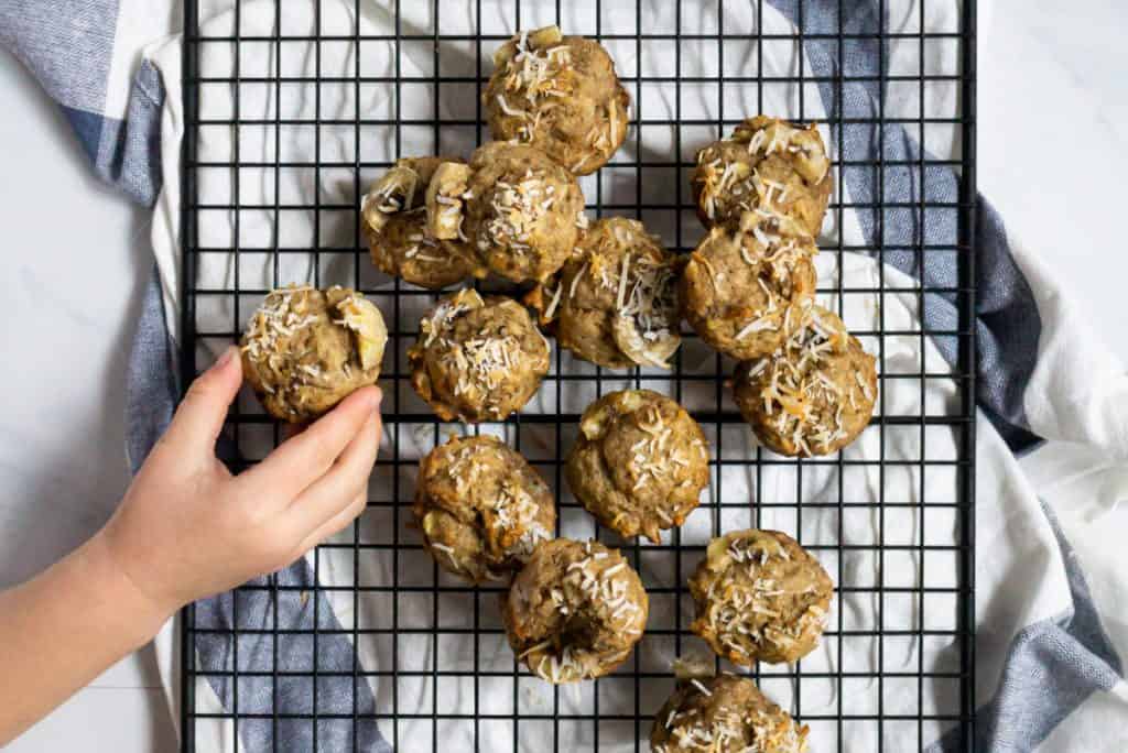 Mini coconut baby banana muffins on a cooling rack with a toddler hand reaching to grab one muffin.