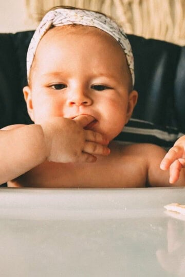Baby sitting in a high chair with a piece of finger food in front of her.