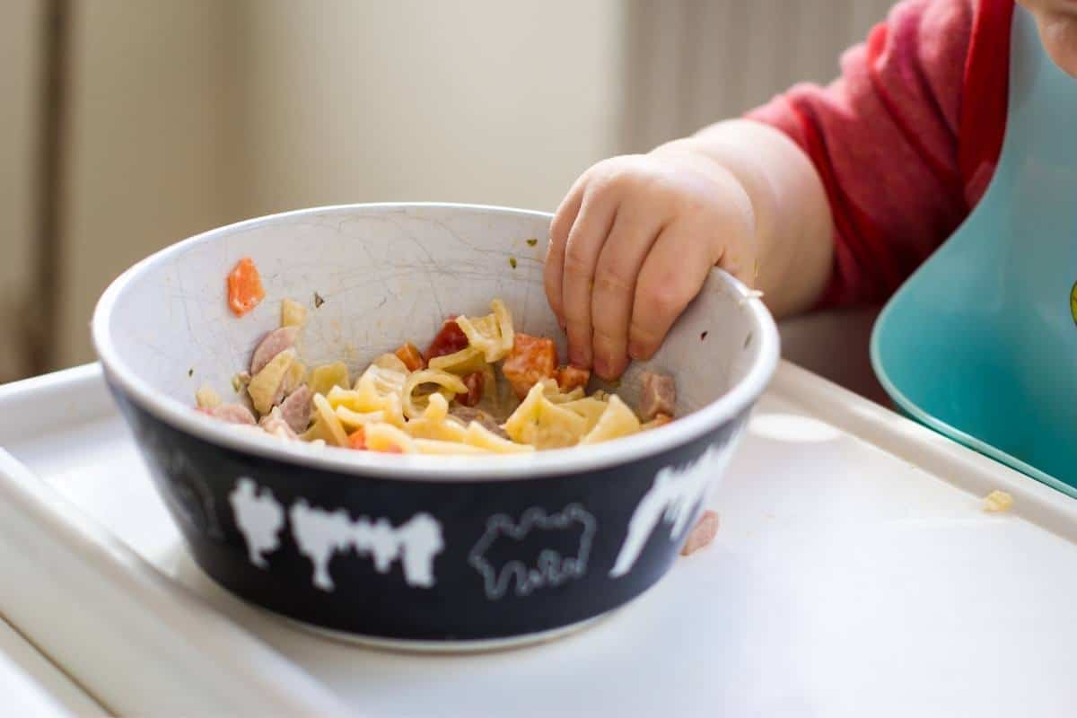 A bowl of noodle soup on a tray with a baby reaching for it