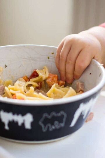 A bowl of noodle soup on a tray with a baby reaching for it