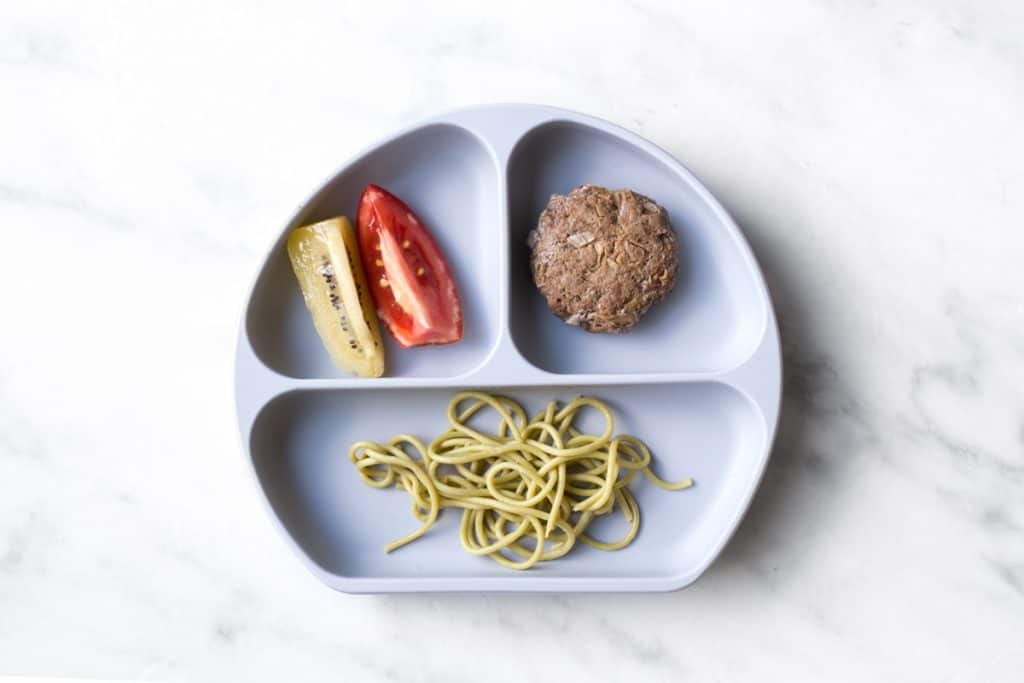 A divided baby plate with spaghetti noodles in one section, a baby beef meatball in another, and a slices of kiwi and a slice of tomato in the last section.