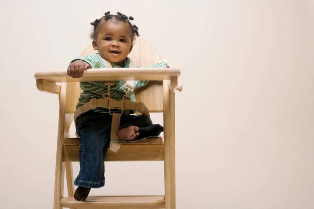 Smiling black baby sitting buckled into a wooden high chair.