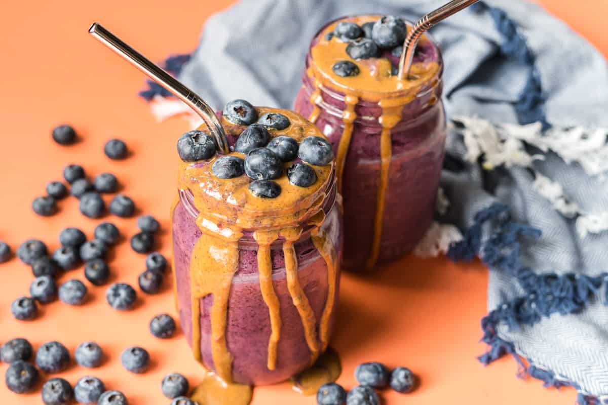 two mason jars with blueberry baby smoothies with almond butter drizzled on the side and loose blueberries surrounding the scene