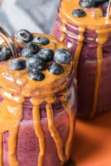 two mason jars with blueberry baby smoothies with almond butter drizzled on the side and loose blueberries surrounding the scene