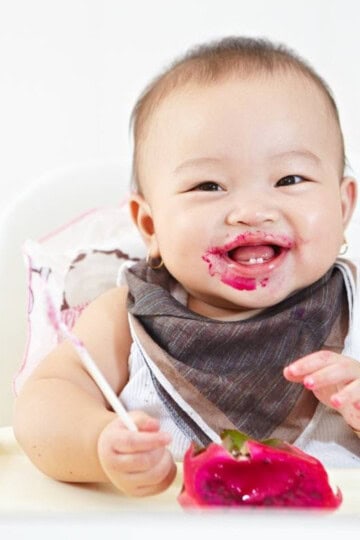 Happy baby wearing a bandana bib, sitting in a high chair, eating dragon fruit