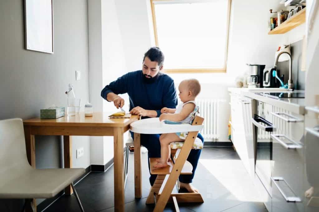 baby sitting in high chair while man, presumably, the father, prepares some food at the table