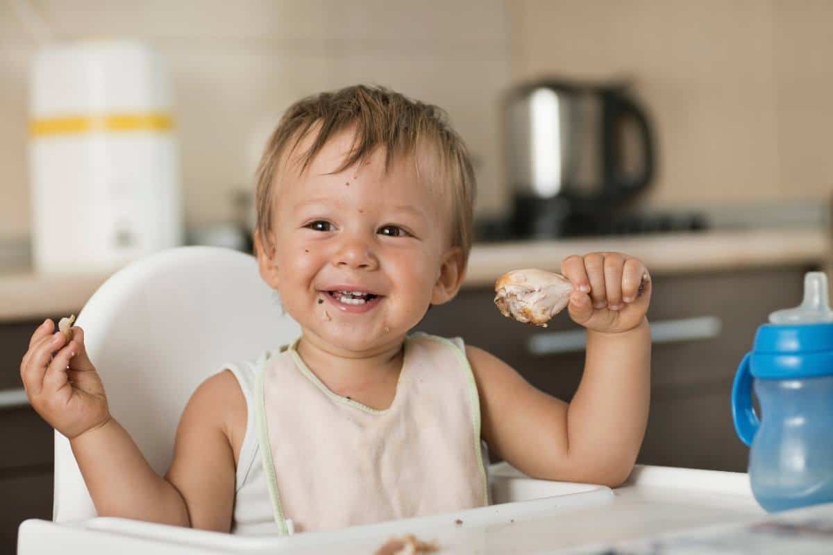 a blond baby sitting in a high chair holding a chicken drumstick