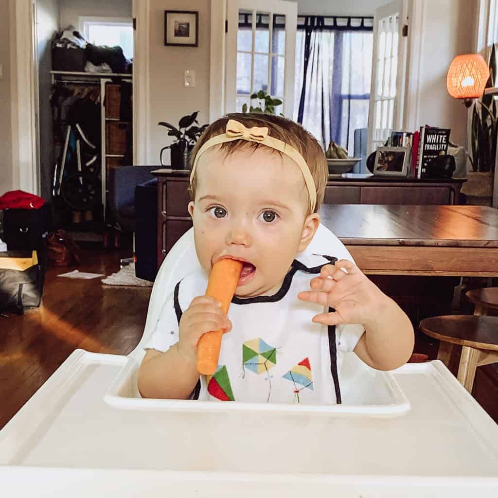 A baby in a high chair gumming on a large raw carrot as a hard munchable.