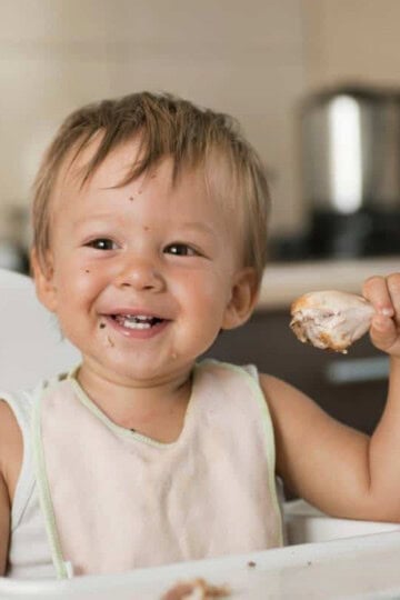 a blond baby sitting in a high chair holding a chicken drumstick