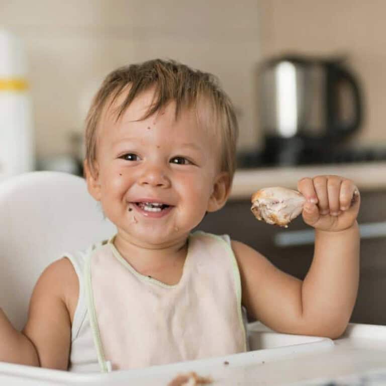 a blond baby sitting in a high chair holding a chicken drumstick