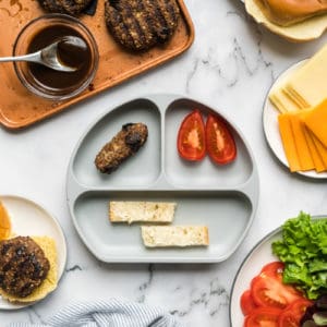 A baby plate with a baby prune burger shaped into a log, tomato wedges, and toast strips, surrounded by the toppings with which an adult would make a burger.