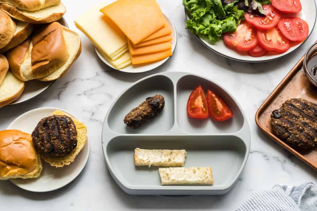 baby plate with baby kofta burger, toast, and tomato strips, surrounded by what the adults are eating: full burgers with the fixin's.