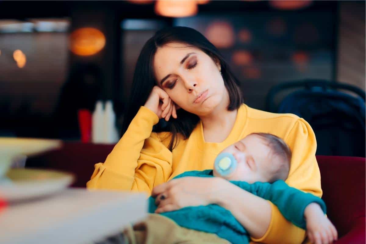 tired mom in a yellow shirt holding a sleeping baby at a table