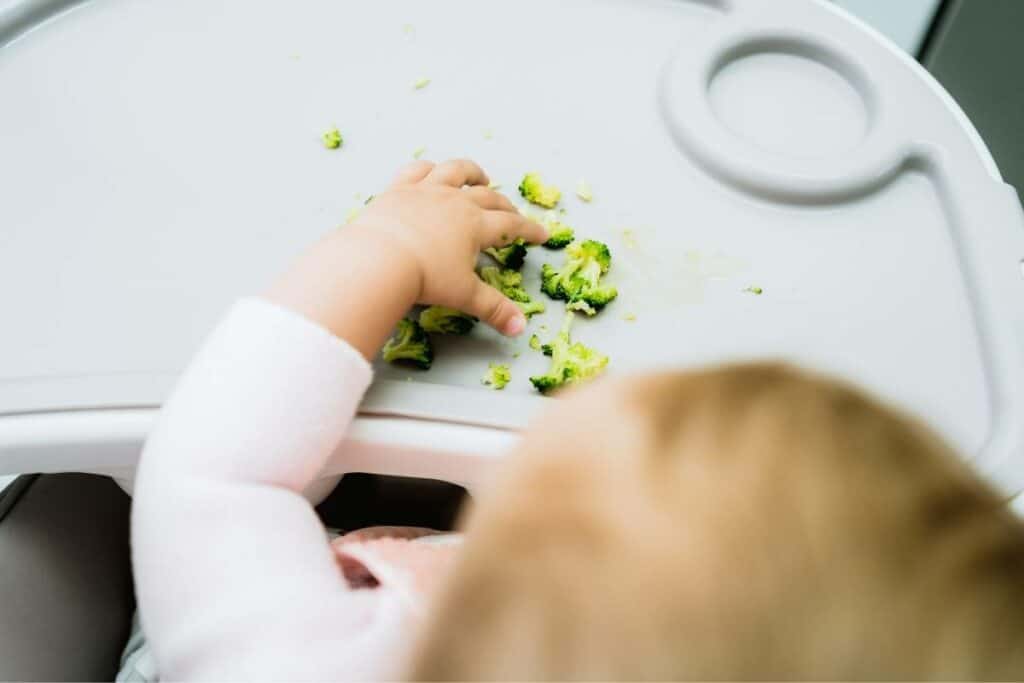 birds eye view of a blond baby in a high chair reaching for small pieces of steamed broccoli