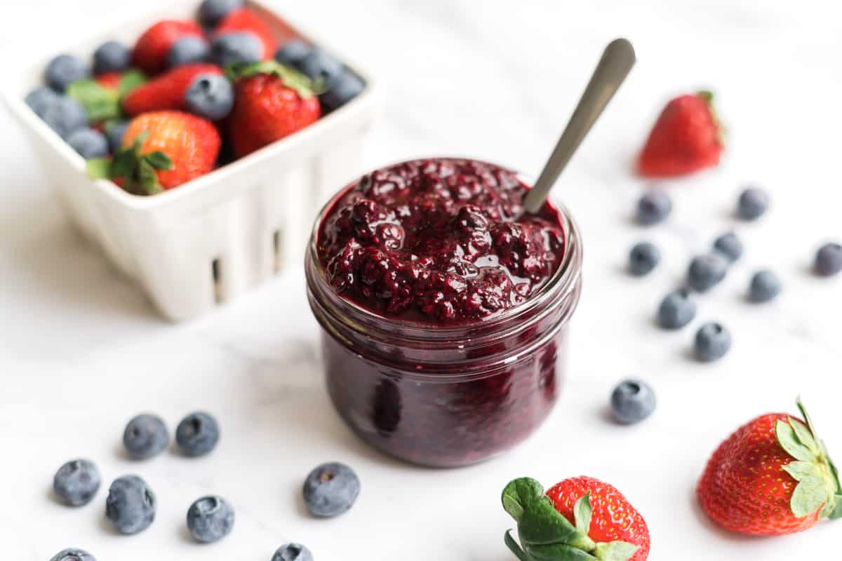 a jar of berry chia sauce surrounded by fresh strawberries and blueberries
