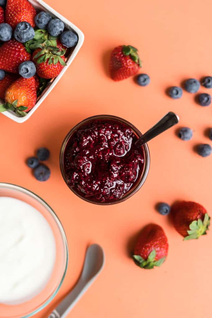 jar of chia berry sauce on pink background beside a bowl of yogurt and fresh berries