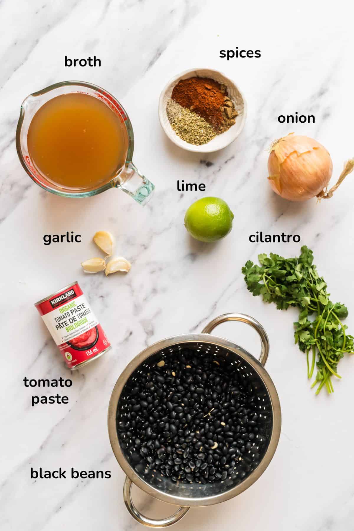 flat lay of ingredients for black bean dip recipe such as broth, spices, onion, lime, cilantro, dried black beans in a colander, tomato paste and garlic. 