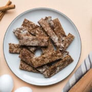 a plate of baby teether biscuits surrounded by cinnamon sticks, a bowl of prunes, eggs, and a rolling pin