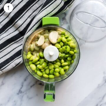 flat lay of ingredients in a food processor: garlic, olive oil, tahini, lemon juice and edamame. 