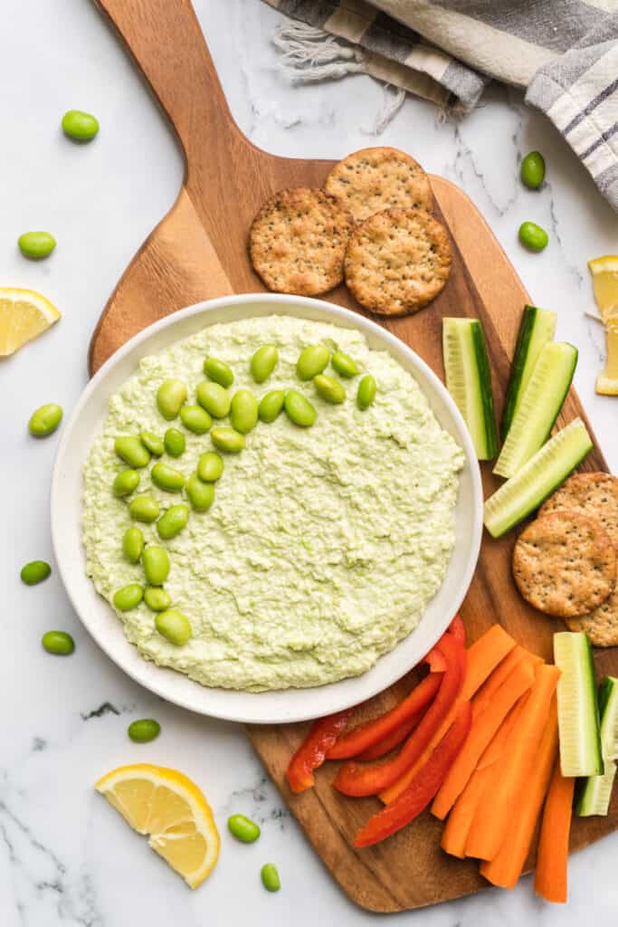 A bowl of edamame baby dip surrounded by cut veggies and crackers.