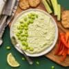 a bowl of creamy edamame hummus surrounded by crackers and veggies on a green background