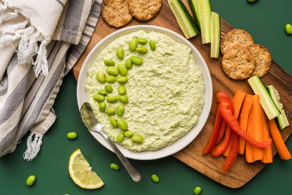 a bowl of creamy edamame hummus surrounded by crackers and veggies on a green background