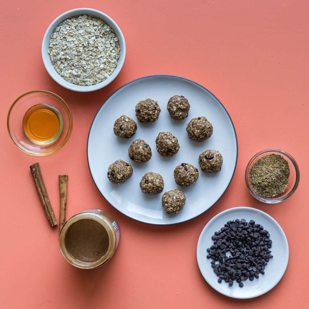 toddler oatmeal ball ingredients on pink background: oats, honey, cinnamon, almond butter, flax, and mini chocolate chips