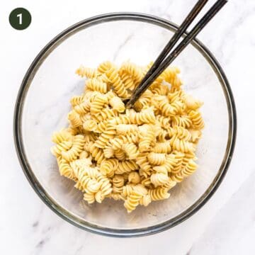 Bowl that contains cooked rotini pasta and salad tossing spoons.