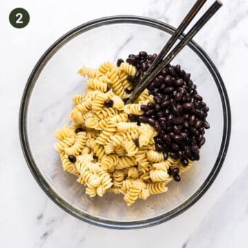 Bowl that contains cooked rotini pasta with canned black beans and salad tossing spoons.