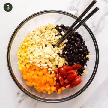 Bowl that contains cooked rotini pasta , black beans, cherry tomatoes, corn, bell pepper and salad tossing spoons.