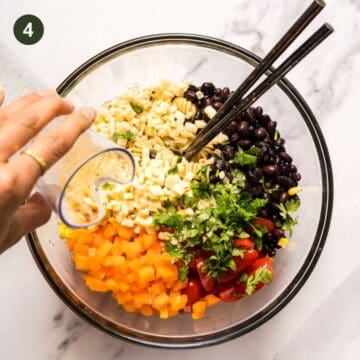 Bowl that contains cooked rotini pasta , black beans, cherry tomatoes, corn, bell pepper and salad tossing spoons. There is fresh cilantro as a garnish and someone pouring dressing into the bowl.