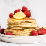 A plate of a stack of fluffy lemon ricotta pancakes with poppyseeds topped with strawberries.