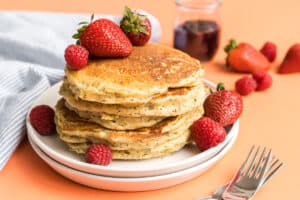A stack of fluffy lemon ricotta pancakes with poppyseeds with a pink background.