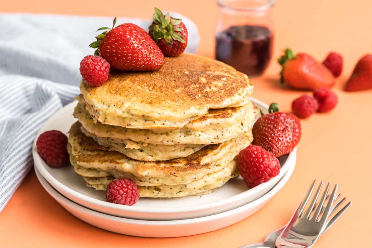 A stack of fluffy lemon ricotta pancakes with poppyseeds with a pink background.