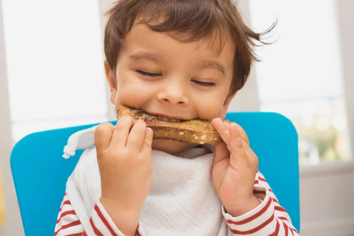 toddler with brown hair at a high chair biting into a slice of toast.
