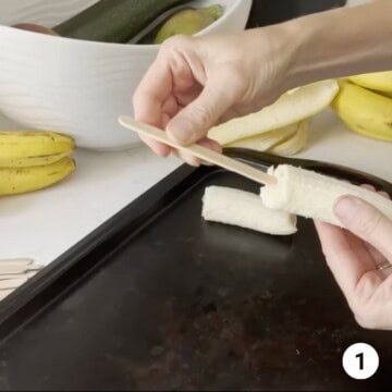 A popsicle stick is being inserted into a banana sliced in half.