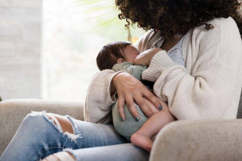 A mother with brown curly hair wearing a white sweater and jeans with a rip in them, nursing a little baby with dark brown hair.