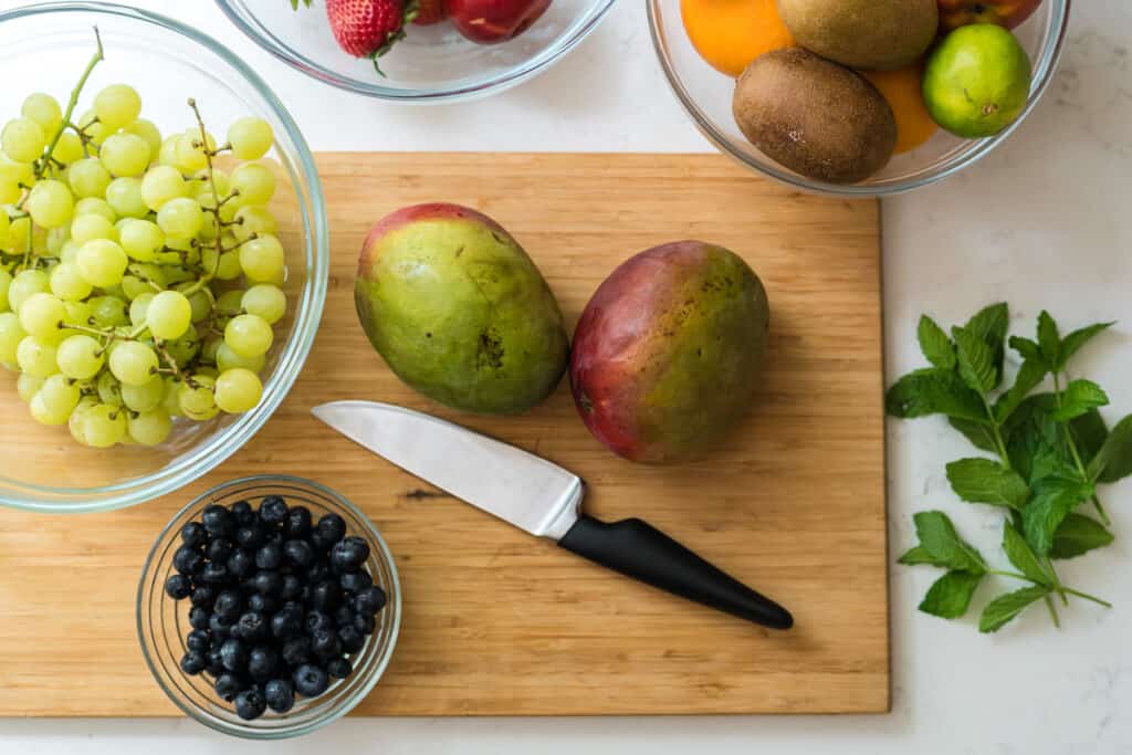 A wooden cutting board with grapes, blueberries, mangoes, strawberries, oranges, kiwi, limes, and a knife