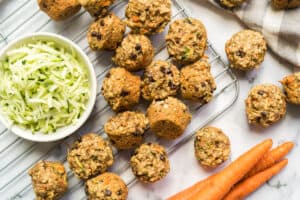 Carrot Zucchini Mini Muffins on a cooling rack with a bowl of shredded zucchini and whole carrots.