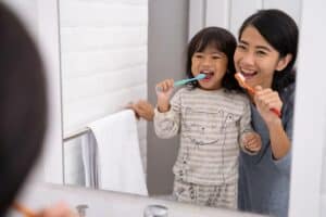 Young girl brushing her teeth in front of the bathroom mirror with her mother leaning over and modeling how to brush teeth.