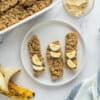 A plate with three pieces of porridge fingers for baby led weaning surrounded by an open banana, a pan of oatmeal fingers, and a small bowl of tahini.