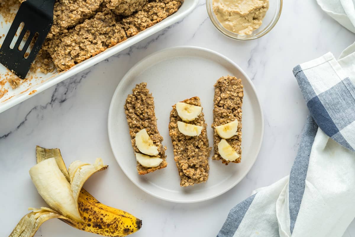 A plate with three pieces of porridge fingers for baby led weaning surrounded by an open banana, a pan of oatmeal fingers, and a small bowl of tahini.