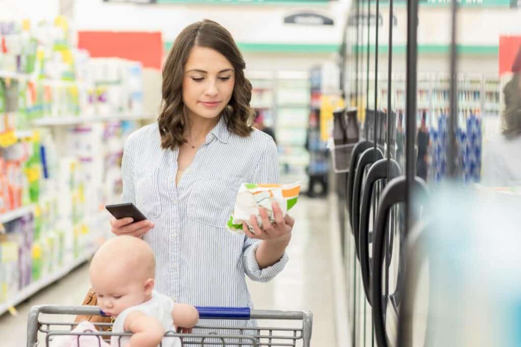 A mother pushing a grocery cart with a baby in the seat, is looking at a food label beside the fridge or freezer section.