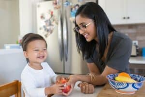 A mother with long black hear wearing glasses is holding an apple for her toddler son to explore. They are both in the kitchen.