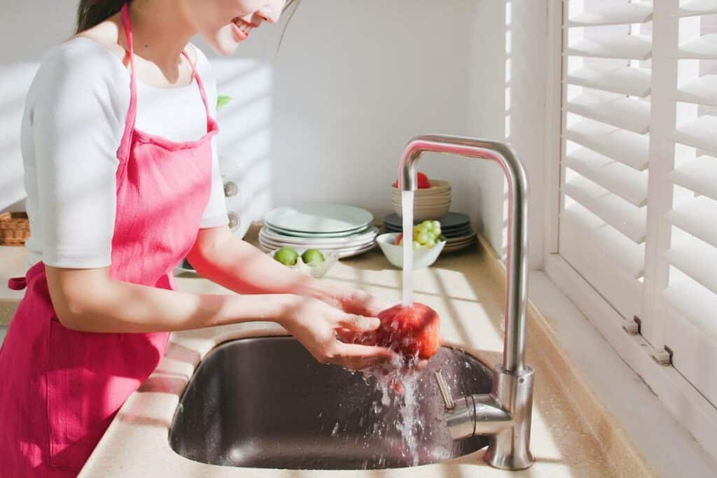 A woman wearing an apron is washing an apple at a kitchen sink.